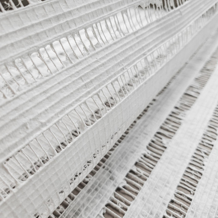Close-up of Handmade Geloma Kozo Paper (White) by Kozo Studio, showcasing layers of textured fabric akin to fine mulberry paper with horizontal lines and irregularly spaced cut-out sections forming a pattern of thin and wider segments. The background displays similar layered patterns.