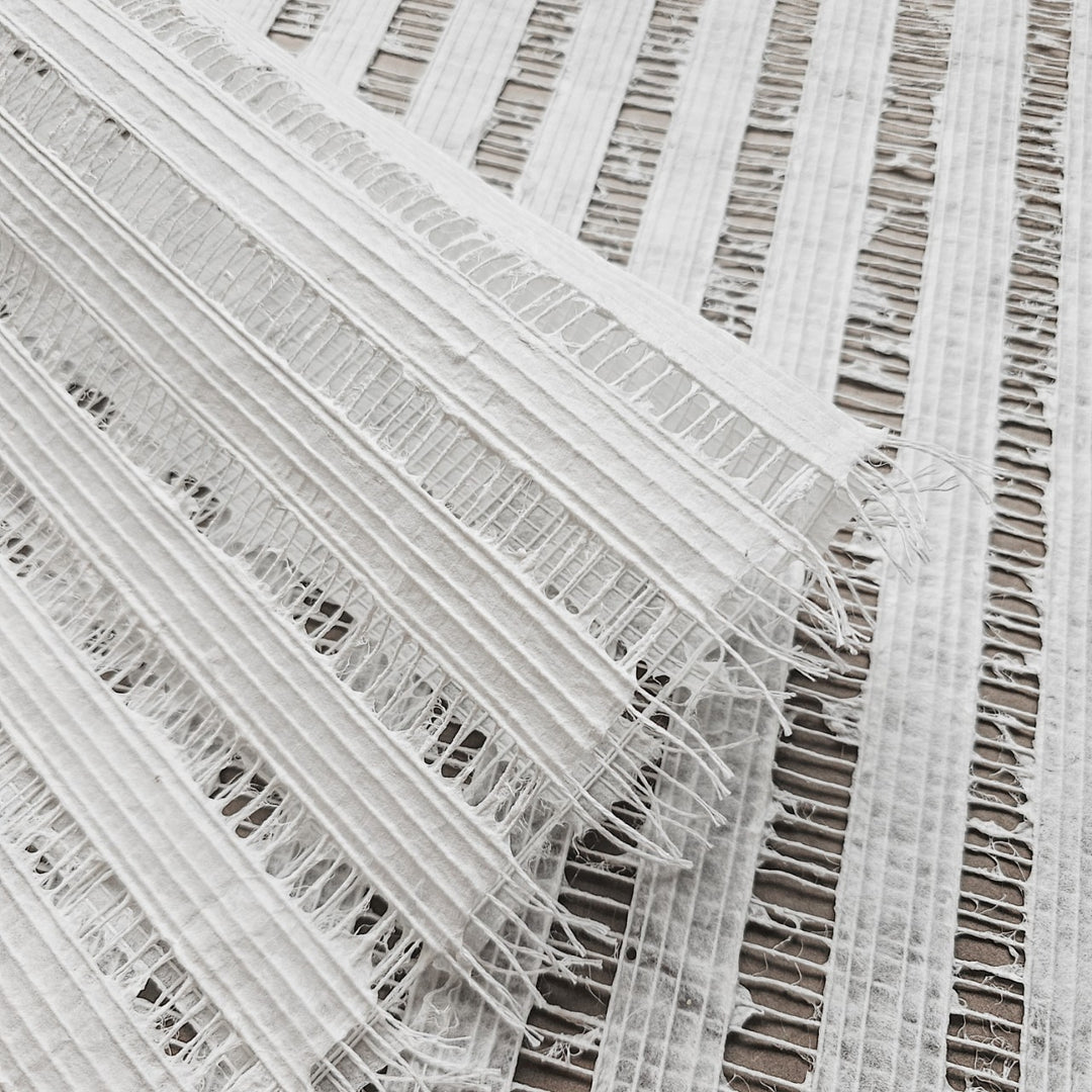 An image showcases the Handmade Geloma Kozo Paper (White) by Kozo Studio, featuring a close-up of textured woven fabric akin to fine rice paper. Displayed diagonally and with frayed edges, it reveals an intricate raised line pattern that adds depth and contrast.