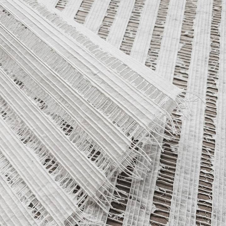 An image showcases the Handmade Geloma Kozo Paper (White) by Kozo Studio, featuring a close-up of textured woven fabric akin to fine rice paper. Displayed diagonally and with frayed edges, it reveals an intricate raised line pattern that adds depth and contrast.