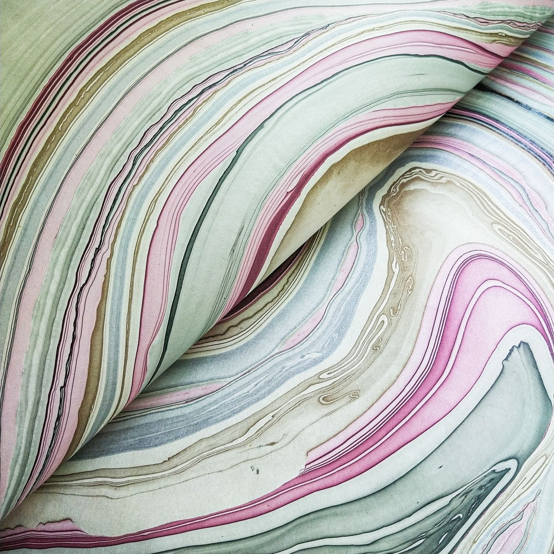 Close-up of an open book reveals Marble Kozo Paper by Kozo Studio, featuring swirling marbled pages in pastel pink, green, and beige. The dynamic abstract design resembles Mulberry Paper textures or natural stone.