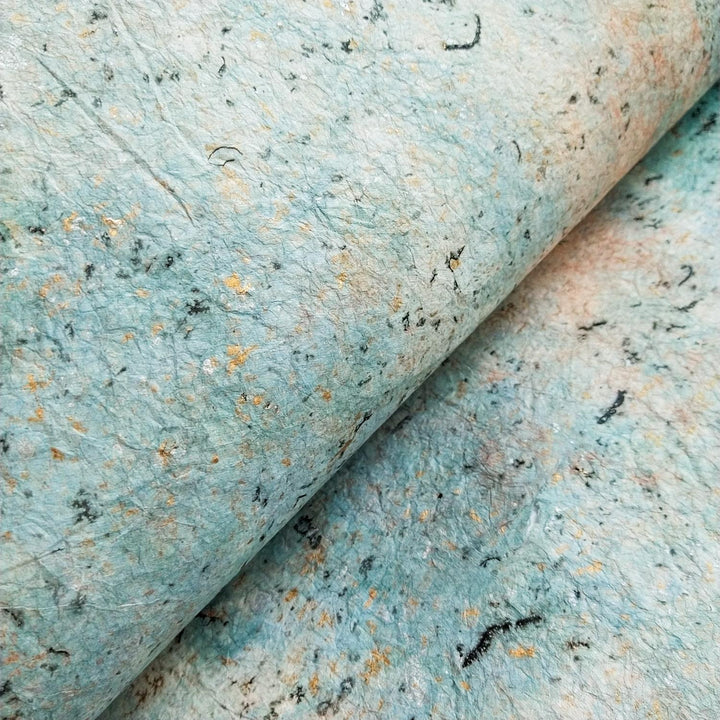 A close-up of Milano Kozo Paper - Design 1 by Kozo Studio reveals a handmade marbled pattern in light blue, teal, and beige. The textured, uneven surface with visible fibers adds an organic, artistic feel.