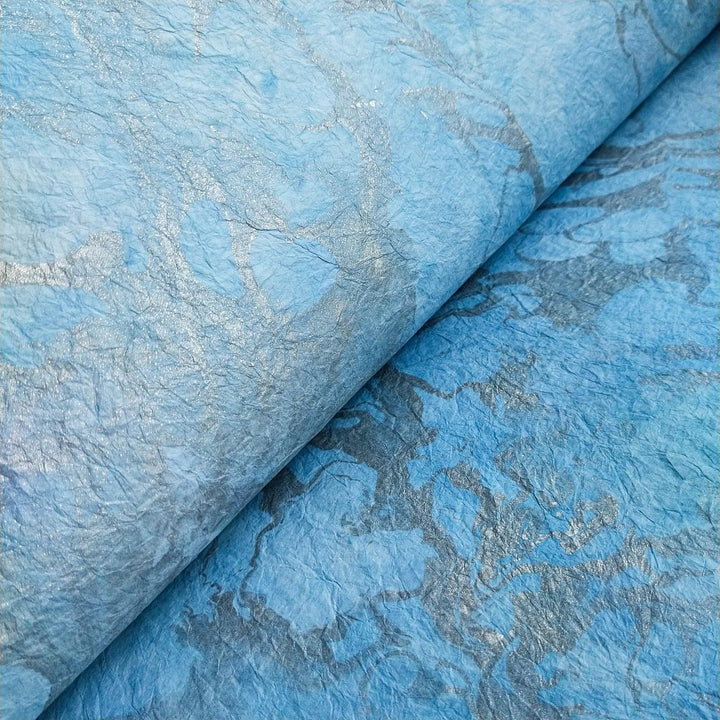 Close-up of Milano Kozo Paper - Design 2 by Kozo Studio, showcasing textured, crumpled mulberry paper with subtle silver marbling. The intricate patterns and folds create a dynamic surface with varying shades of blue and hints of metallic sheen.