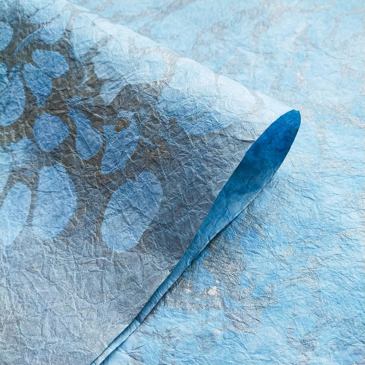 The close-up of Milano Kozo Paper - Design 2 by Kozo Studio reveals its textured blue paper with a curved edge, featuring abstract marbled dark spots and lighter areas for an organic blend of blue tones.
