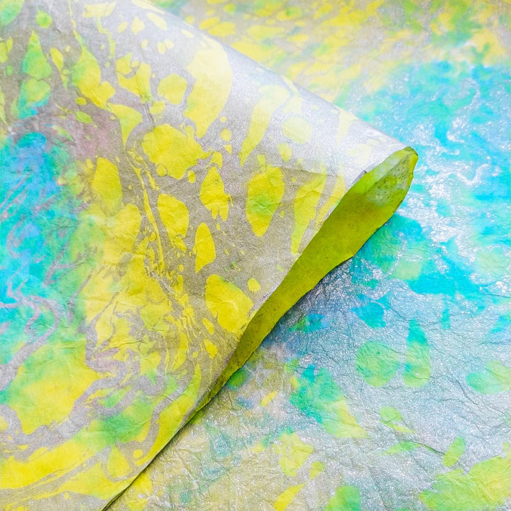 Close-up of Milano Kozo Paper - Design 3 by Kozo Studio, featuring textured mulberry paper with intricate abstract patterns in vibrant green, yellow, and blue. The image shows a slightly curled edge revealing its colorful design.