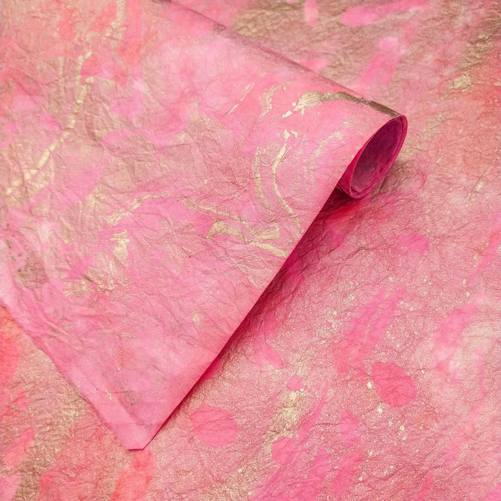 Close-up of a rolled piece of Milano Kozo Paper - Design 4 by Kozo Studio, showcasing a pink and gold textured design. The paper appears crinkled with abstract patterns, blending the vibrant colors for a shimmering effect.