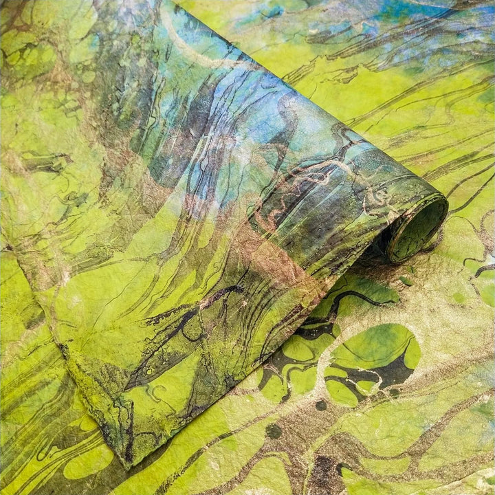 Close-up of Milano Kozo Paper - Design 5 by Kozo Studio features a vibrant green marbled texture with swirls of black and various shades of green. It has a smooth, glossy surface and intricate, fluid patterns. A curled edge reveals more design details.