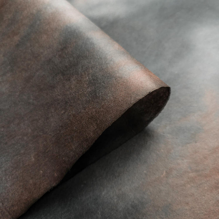 A close-up of Ombre Kozo Paper (#10 Midnight Umber) by Kozo Studio, featuring a textured, weathered brown mulberry appearance with a rolled corner on a flat surface. This papers mottled design gives it a rustic look.