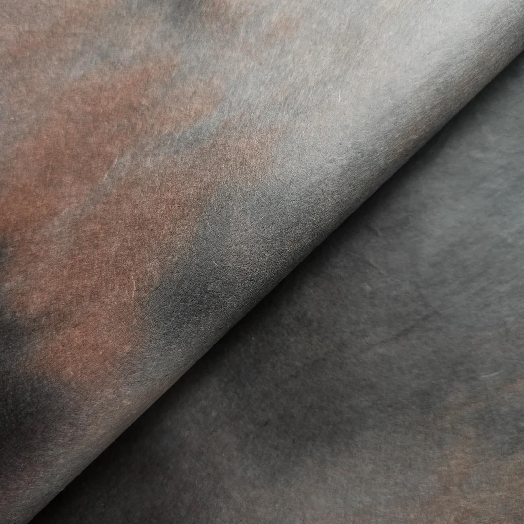 A close-up of Ombre Kozo Paper (#10 Midnight Umber) by Kozo Studio reveals its smooth mulberry texture with dark browns and subtle grays. Soft lighting enhances its depth and contrast, creating a shadowed effect.