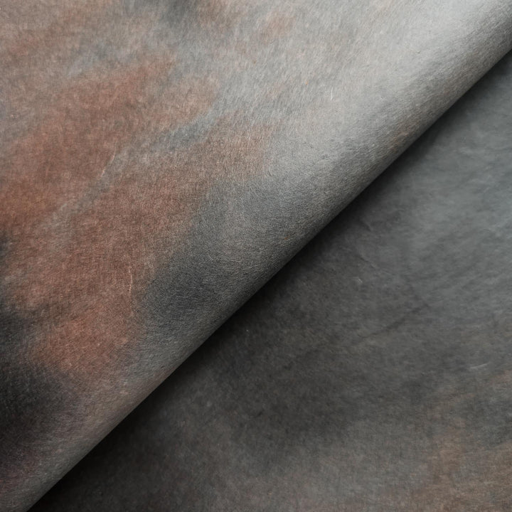 A close-up of Ombre Kozo Paper (#10 Midnight Umber) by Kozo Studio reveals its smooth mulberry texture with dark browns and subtle grays. Soft lighting enhances its depth and contrast, creating a shadowed effect.