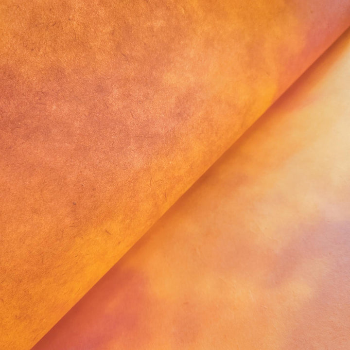 A close-up of Ombre Kozo Paper (#7 Amber Dawn) from Kozo Studio shows two overlapping sheets with a gradient from orange to light orange. The smooth textured mulberry creates soft shadows enhancing color transitions.
