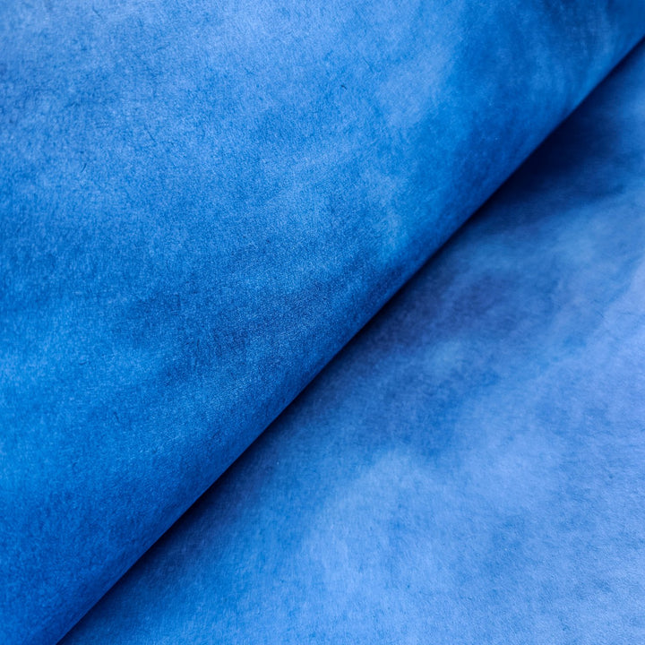 A close-up of Ombre Kozo Paper (#8 Cerulean Blue) from Kozo Studio reveals its textured surface with subtle tonal variations of mulberry paper. It has a soft, velvety look, showcasing thickness and rich color as its rolled in layers.
