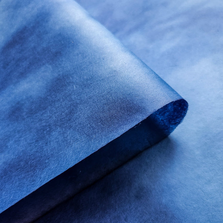 A close-up of Ombre Kozo Paper (#8 Cerulean Blue) by Kozo Studio shows its semi-transparent texture with a curled corner casting a shadow. The faint texture is visible, complemented by a matching background, enhancing the monochromatic theme.