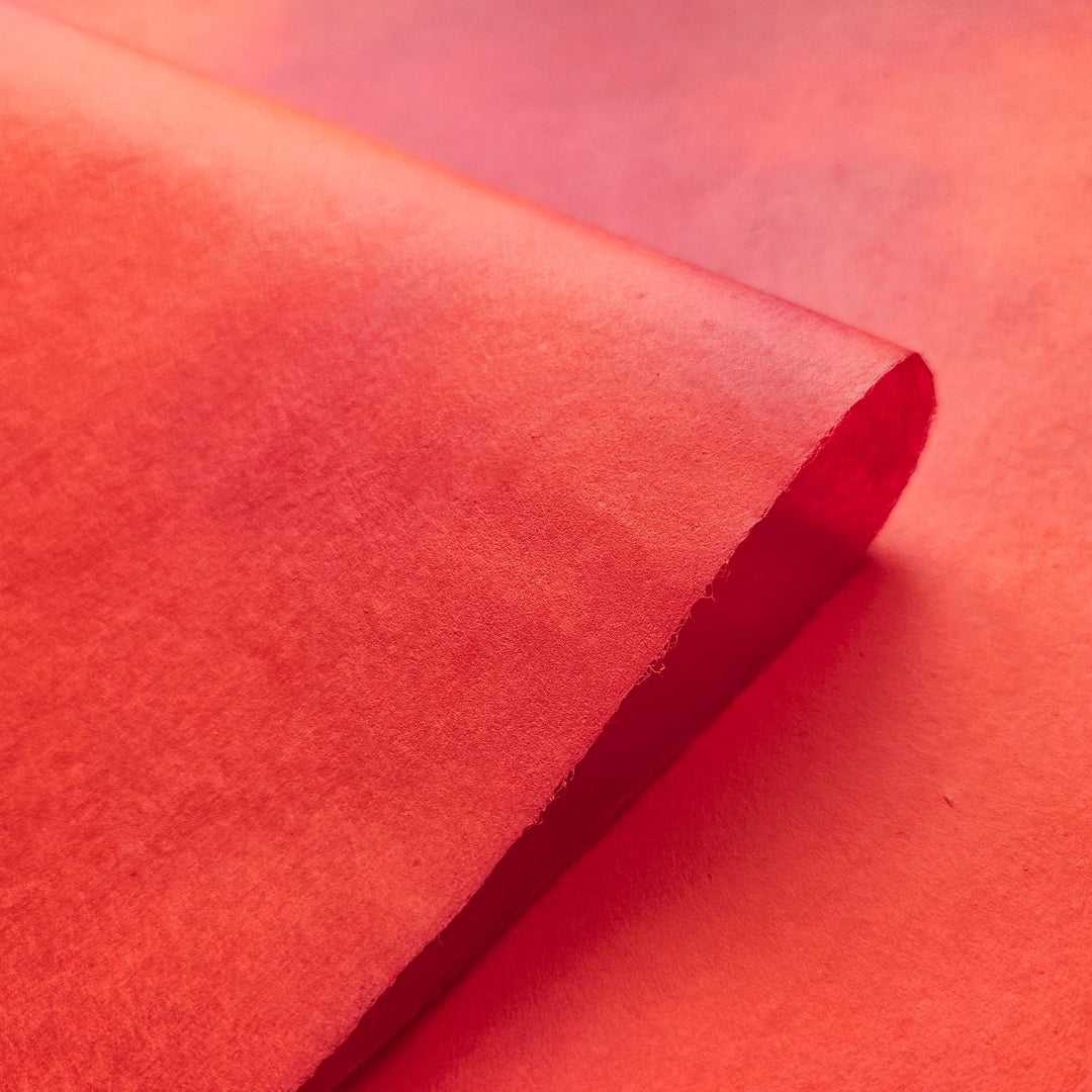 A close-up of Kozo Studios Ombre Kozo Paper (#9 Scarlet Dusk) with a slightly curled edge, set against a smooth red background. It has soft texture of mulberry paper, and the lighting highlights its warm, vibrant tone.