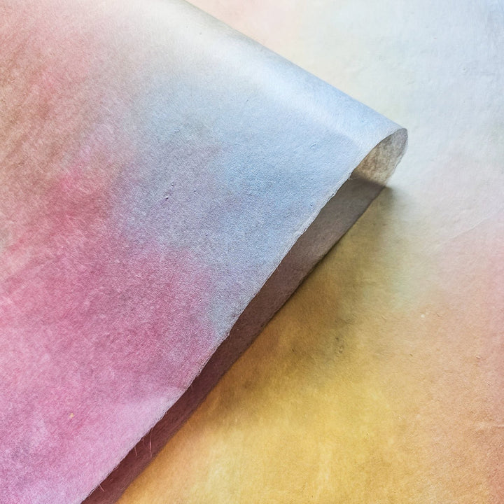A roll of Kozo Studio Ombre mulberry paper in soothing pastel tones, blending yellow and peachy pink hues with a handmade paper finish ideal for crafts and decor.