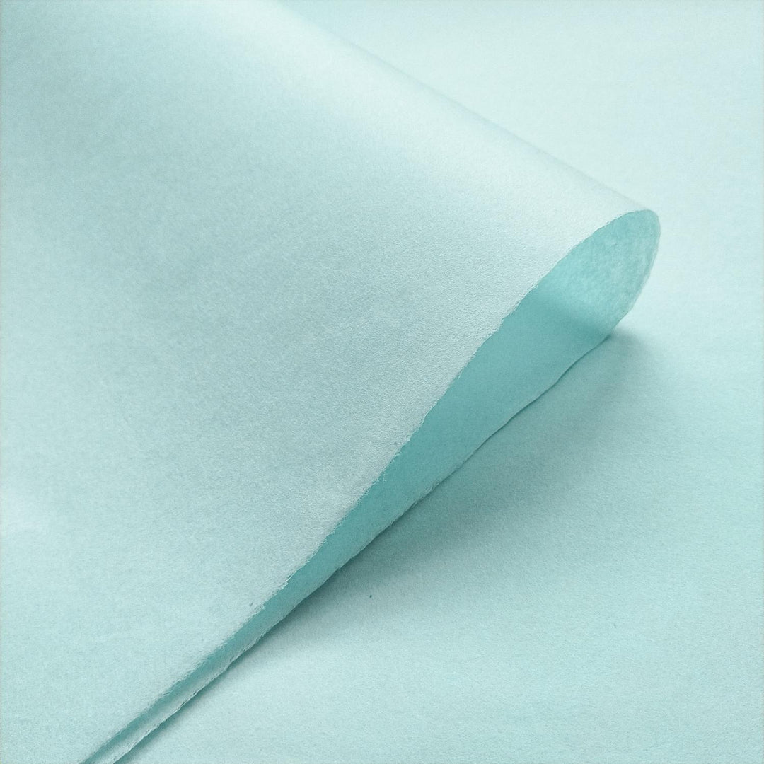Handmade mulberry paper in sky blue by Kozo Studio, displayed with a diagonal crease to emphasize its fine texture and light-reflecting surface.