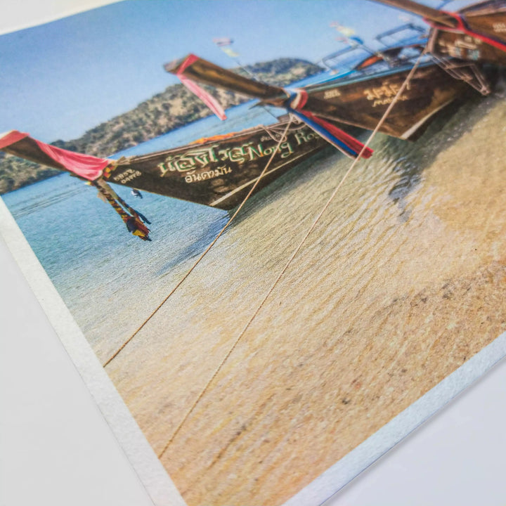 Traditional longtail boats with colorful fabric at their prows are docked on a sandy beach, reminiscent of the artistry in Kozo Studios A4 Thin Natural Kozo Paper. The clear, calm water and distant hills under a blue sky enhance this vibrant scene.