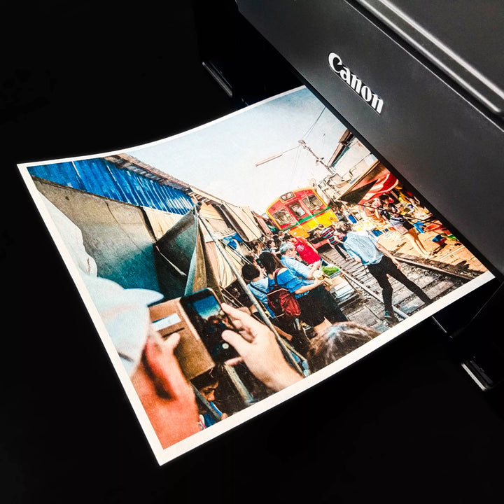A Canon printer is producing a vibrant photo on A4 White Kozo Paper by Kozo Studio. The image shows a busy outdoor market with colorful stalls and a passing train, while two people photograph the lively moment on their phones, preserving it on this special medium.