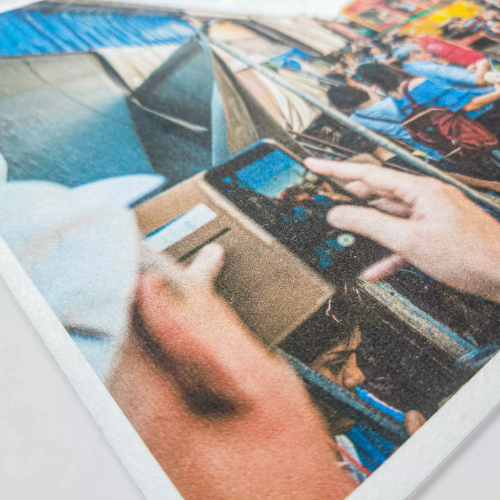 At a lively outdoor market, a cap-wearing individual snaps a photo with their smartphone amidst bustling activity. People interact with vibrant stalls featuring Kozo Studios colorful tents and booths adorned with A4 White Kozo Paper, capturing the essence of creativity and engagement.