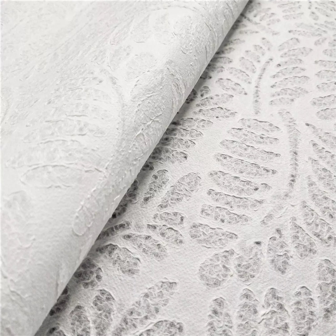 Close-up of textured white Handmade Lace Kozo Paper (Fern) by Kozo Studio. This is a mulberry paper with an embossed fern-like pattern. The partially folded paper casts a soft shadow, highlighting the intricate details of its raised design.