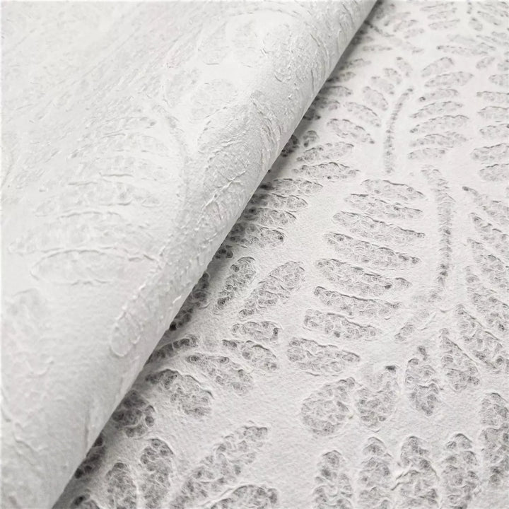 Close-up of textured white Handmade Lace Kozo Paper (Fern) by Kozo Studio. This is a mulberry paper with an embossed fern-like pattern. The partially folded paper casts a soft shadow, highlighting the intricate details of its raised design.