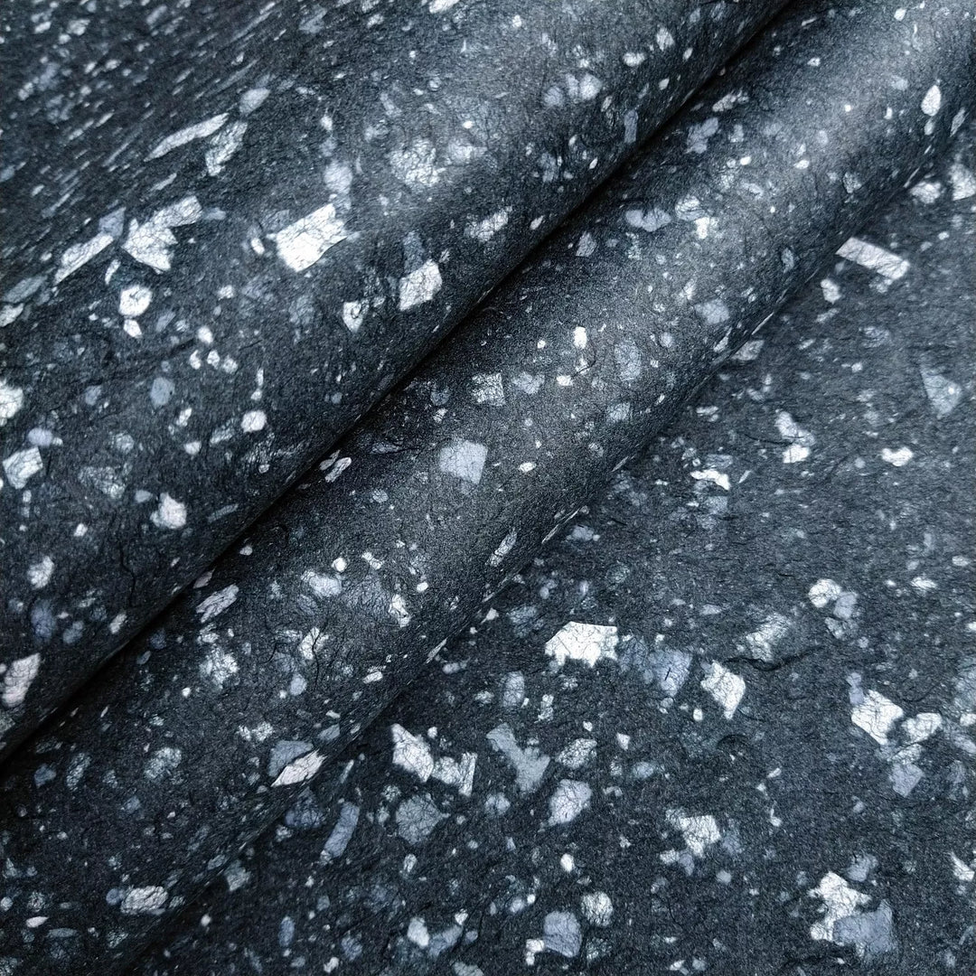 Close-up of Granite Kozo Paper (Midnight) by Kozo Studio, featuring a dark fabric-like texture with irregular white and light gray specks. The smooth surface has a subtle sheen, reminiscent of a starry night sky on Mulberry Paper.