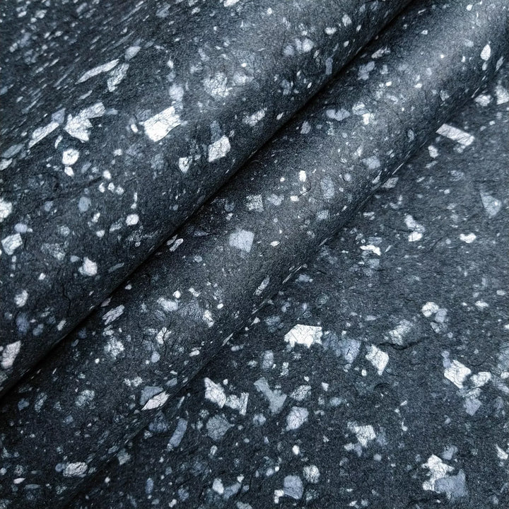 Close-up of Granite Kozo Paper (Midnight) by Kozo Studio, featuring a dark fabric-like texture with irregular white and light gray specks. The smooth surface has a subtle sheen, reminiscent of a starry night sky on Mulberry Paper.