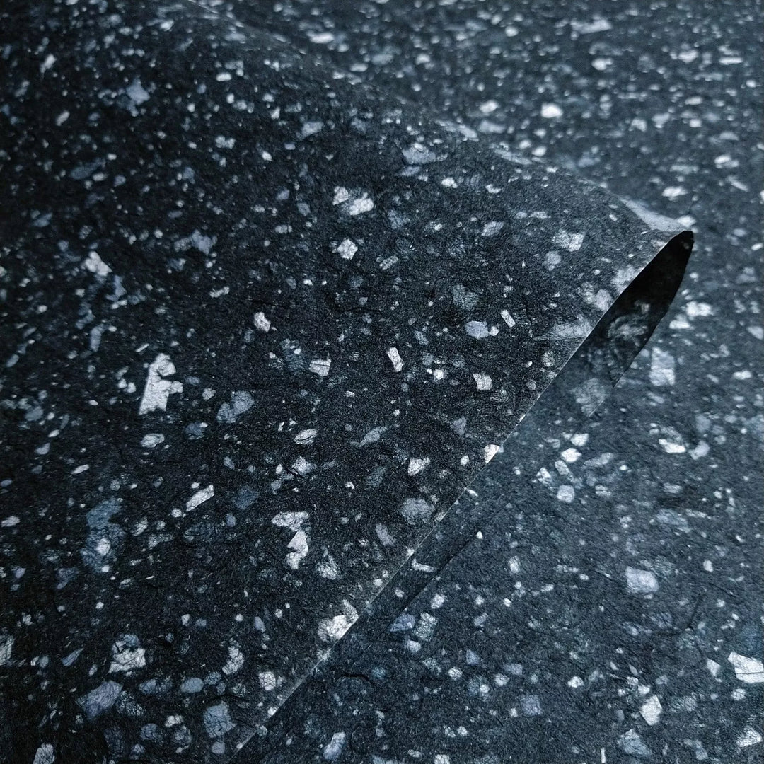 A close-up of Kozo Studios Granite Kozo Paper (Midnight) features a textured dark gray surface with light gray speckles, mimicking stone or terrazzo, and includes a gentle curve of the delicate and organic texture of Mulberry Paper.