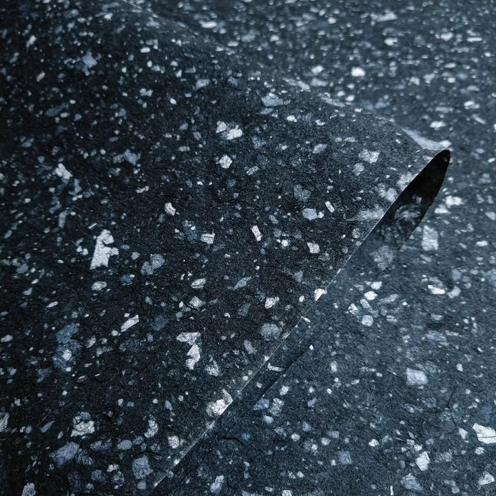 A close-up of Kozo Studios Granite Kozo Paper (Midnight) features a textured dark gray surface with light gray speckles, mimicking stone or terrazzo, and includes a gentle curve of the delicate and organic texture of Mulberry Paper.