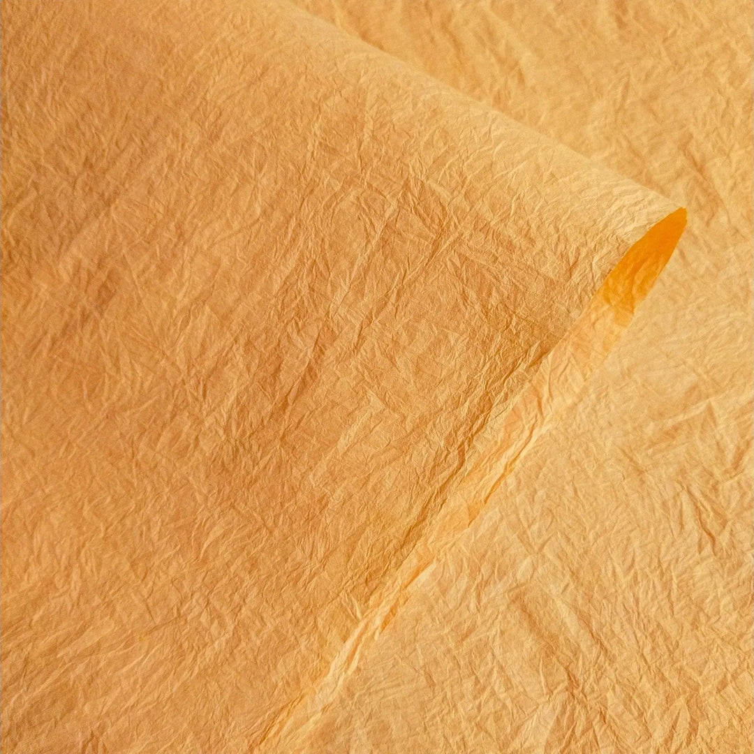 A close-up of a textured Momigami Color Kozo Paper (Yellow) by Kozo Studio, with a slightly curled edge, highlights detailed creases and an uneven surface. The lighting enhances the shades and textures of a finely crafted mulberry paper.