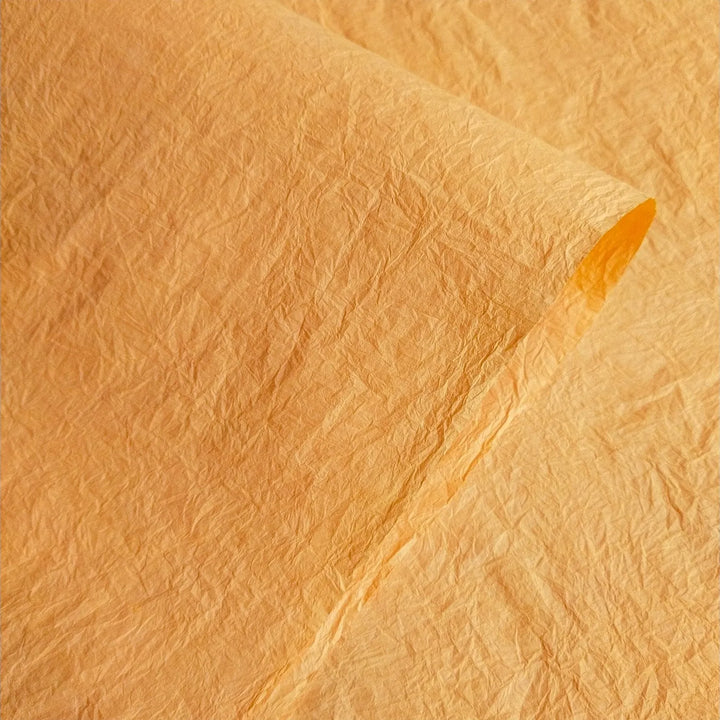 A close-up of a textured Momigami Color Kozo Paper (Yellow) by Kozo Studio, with a slightly curled edge, highlights detailed creases and an uneven surface. The lighting enhances the shades and textures of a finely crafted mulberry paper.