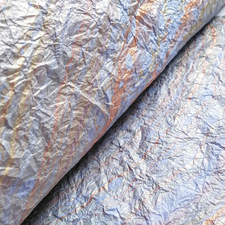 A close-up of the Milano Kozo Paper - Design 13 (Light Blue) by Kozo Studio reveals its textured, iridescent surface. The blend of silver, gold, blue, and purple creates a shimmering, abstract effect reminiscent of crumpled foil or delicate fabric.