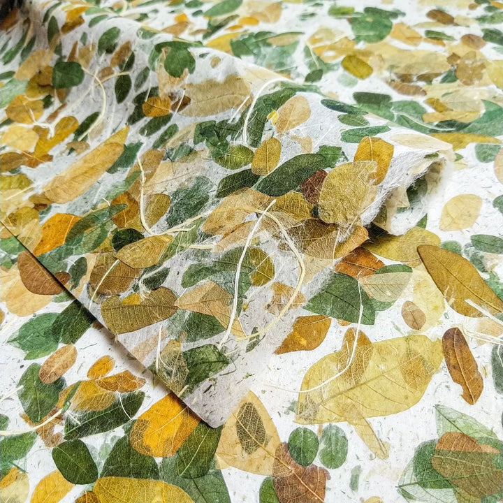 The Very Raintree Kozo Paper by Kozo Studio features a textured mulberry surface adorned with green, yellow, and brown leaf patterns for a natural look, with one edge slightly curled upwards on a flat surface.
