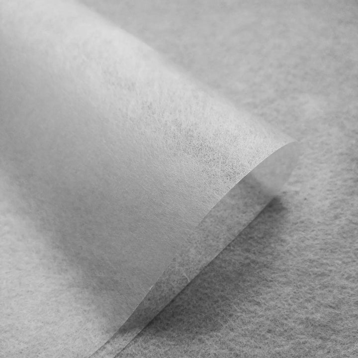 A close-up of a single translucent Conservator No.1 sheet (8.5 gsm) by Kozo Studio, slightly curled at the edge and resting on a similarly textured surface. Comes in packs of 15 sheets.