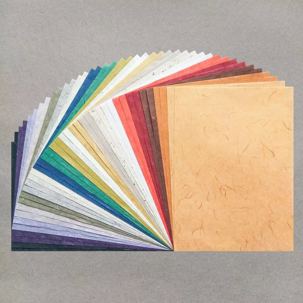 An assortment of A4 Earth Unryu Kozo Paper Set 40 Sheets by Kozo Studio features a fan-like spread of multicolored textured mulberry paper sheets on a gray surface, showcasing hues from purple to orange.