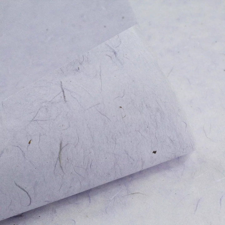 Close-up of Kozo Studios Water-resistant Kozo Paper in Lavender, showcasing its textured finish with visible fibers and specks. The off-white surface appears slightly rough, with a corner turned up to reveal the layer beneath.