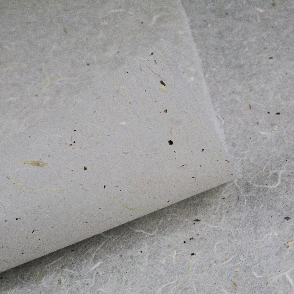 A close-up of water-resistant Kozo Paper from Kozo Studio shows a textured, handmade sheet with visible fibers and small specks curled at one corner atop a similar sheet, boasting a natural, rustic appearance and grayish tone.