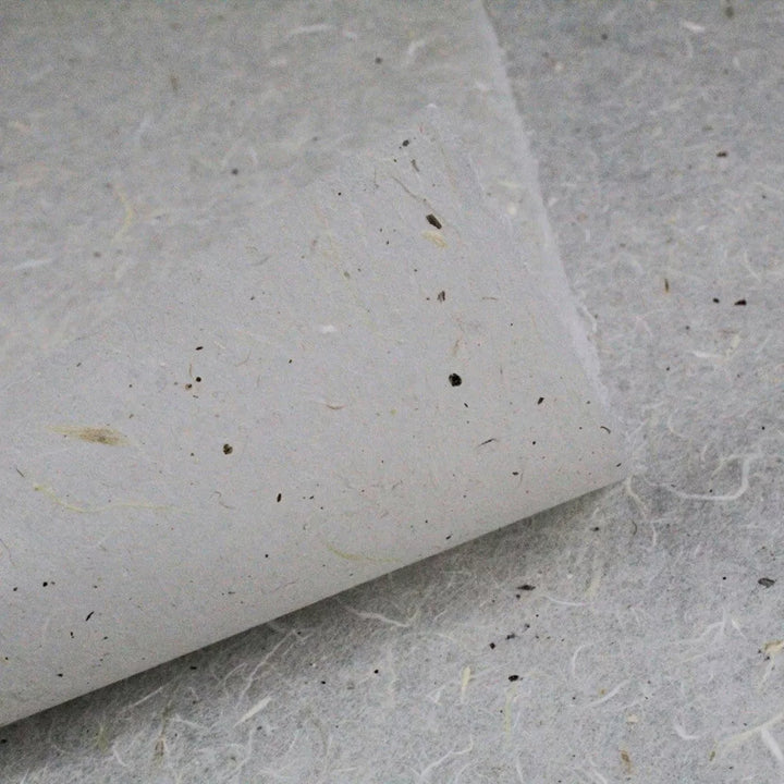 A close-up of water-resistant Kozo Paper from Kozo Studio shows a textured, handmade sheet with visible fibers and small specks curled at one corner atop a similar sheet, boasting a natural, rustic appearance and grayish tone.