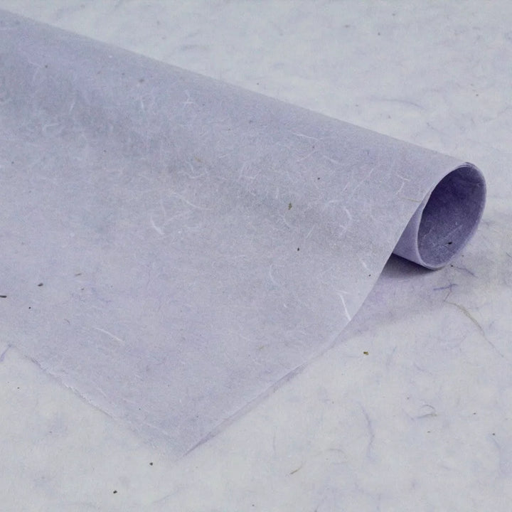A sheet of thin, translucent Water-resistant Kozo Paper (Lavender) by Kozo Studio is rolled at one edge and rests on a similar background, showcasing a subtle texture with visible fibers.