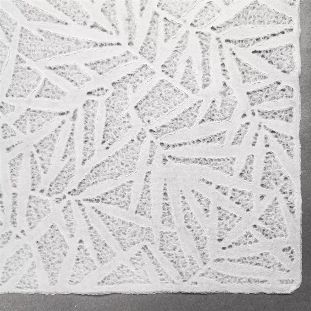 The Handmade Lace Kozo Paper by Kozo Studio features intricate white lace with abstract geometric patterns like triangles and lines, evoking the delicate texture of rice paper against a gray background.