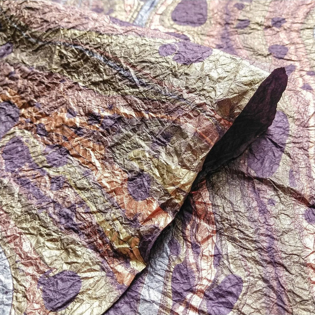 A close-up of Milano Kozo Mulberry Paper - Design 12 (Purple) by Kozo Studio shows crumpled, metallic-textured mulberry paper. Shades of gold and purple create irregular patterns, blending organic creases with circular designs for a dynamic abstract look.