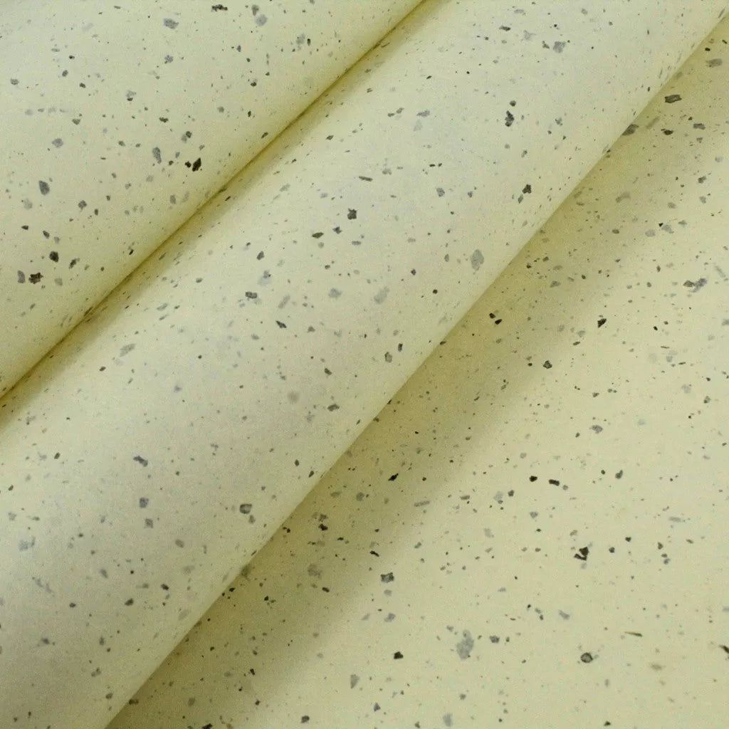 Close-up of handmade paper from Kozo Studio featuring overlapping sheets of pale yellow mulberry paper with speckled kozo leaf confetti, creating soft shadows and delicate texture.