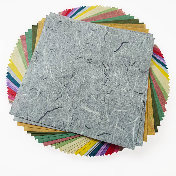 The Origami Unryu Kozo Paper Pack by Kozo Studio features 72 sheets of 6x6 inch square paper in assorted colors such as blue, red, yellow, and green. A gray mulberry marbled-textured sheet is on top, with all papers elegantly fanned out in a circle.