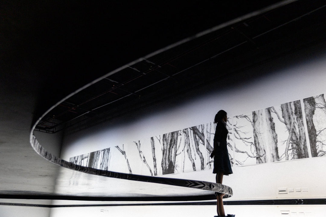 A silhouetted woman stands on a circular platform in a dimly lit exhibition space, observing a series of large black-and-white tree drawings mounted on the wall. The curved structure and minimal lighting create a calm, contemplative atmosphere.