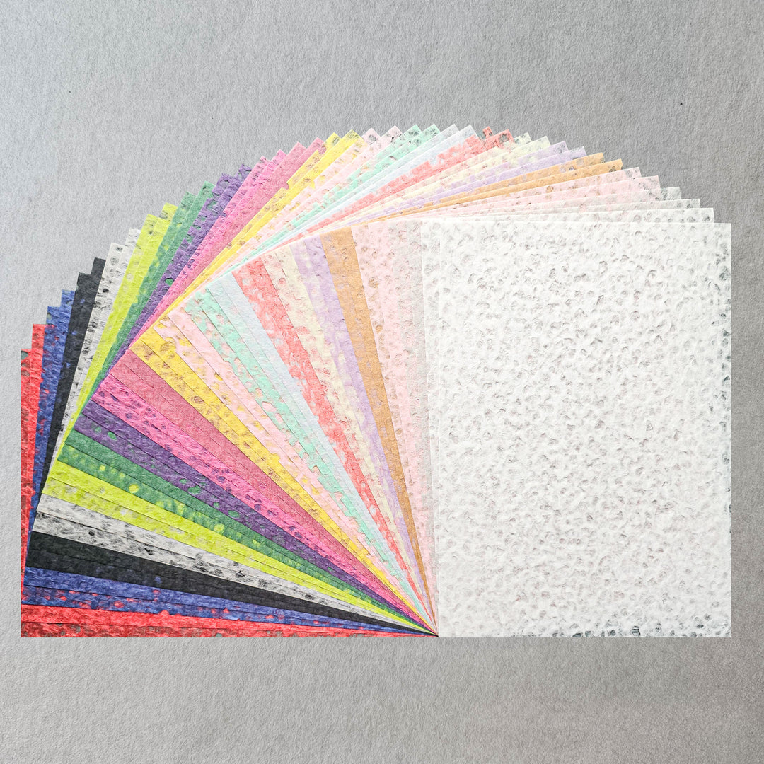 The Kozo Studio Assorted A4 Rakusui Mulberry Paper Set (40 Sheets) features colorful textured sheets fanned out on a gray surface, with a white sheet shown fully—ideal for decorative craft projects.