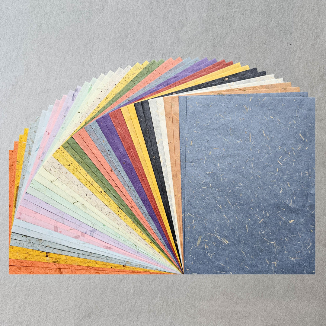 A fan-shaped display of Kozo Studios Assorted A4 Mix Leaf & Flower Mulberry Paper Set (40 Sheets) is spread out on a gray background, with a blue sheet flat on the right—ideal for creative DIY crafts and projects.