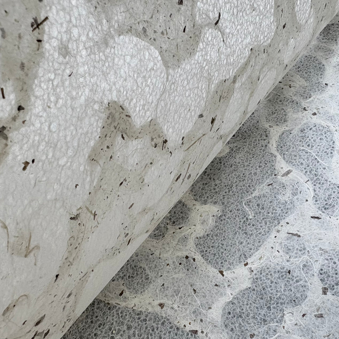 A close-up of Tidal Lace Kozo Paper (Chiri) by Kozo Studio shows its eco-friendly, handmade texture with organic fibers and small dark specks, partially rolled atop a similar fibrous surface.