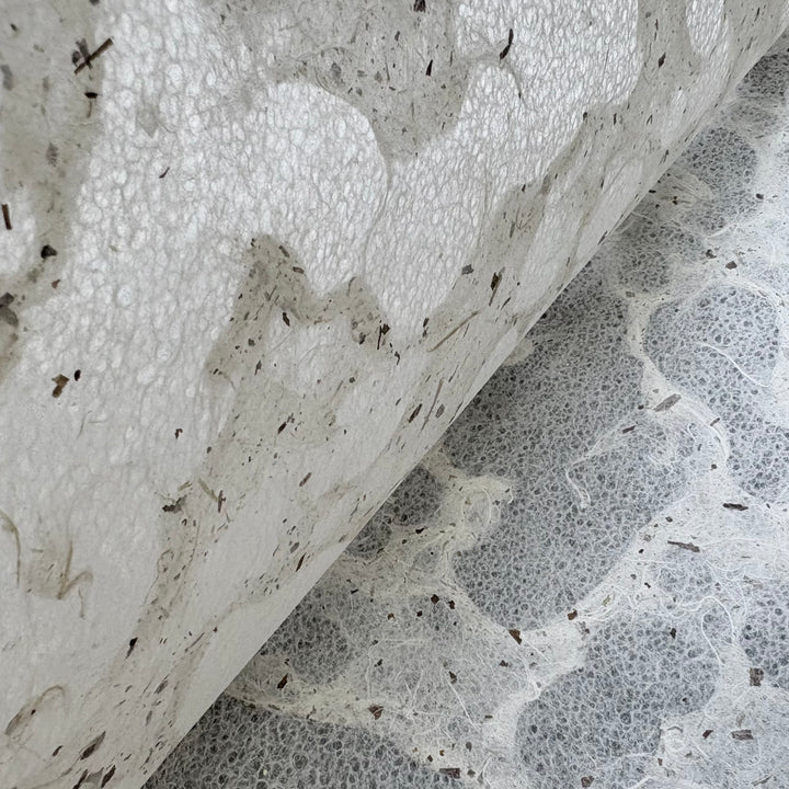 A close-up of Tidal Lace Kozo Paper (Chiri) by Kozo Studio shows its eco-friendly, handmade texture with organic fibers and small dark specks, partially rolled atop a similar fibrous surface.