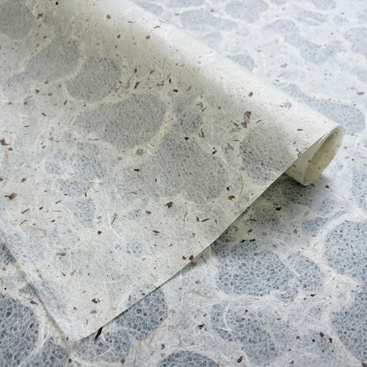 A close-up of Tidal Lace Kozo Paper (Chiri) by Kozo Studio—semi-translucent, eco-friendly handmade mulberry paper with visible plant fibers and brown flecks, partially rolled on a patterned surface.