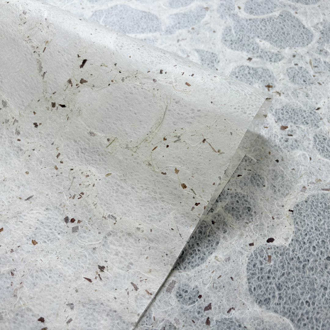 A close-up of Kozo Studio’s eco-friendly Tidal Lace Kozo Paper (Chiri), showing its translucent, handmade texture with visible plant fibers and specks. One sheet is slightly lifted to reveal delicate layers against a matching background.