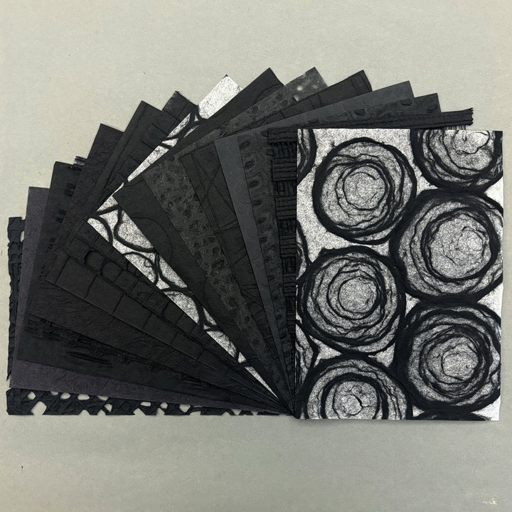 A fan of black textured paper sheets with various patterns, including a prominent black and white circular swirl design on the top sheet, displayed on a neutral gray background.